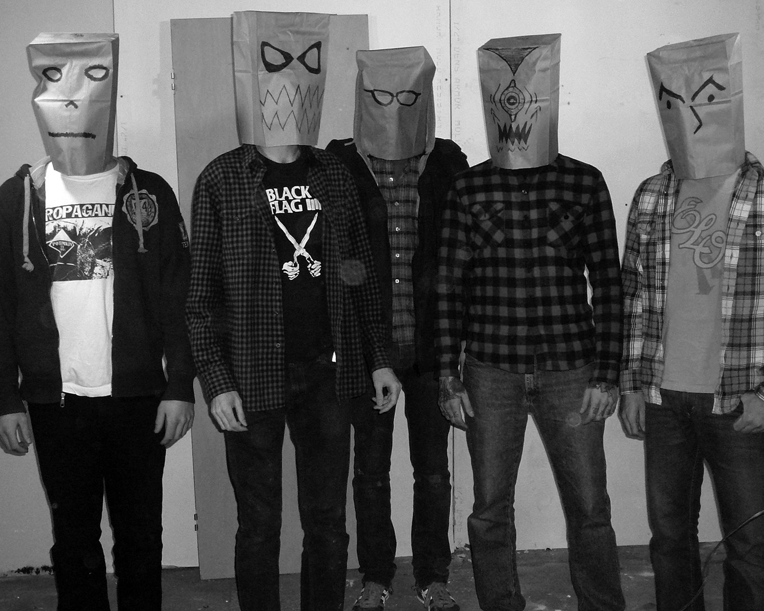 The members of the band standing in an empty room with paper bags on their heads with odd faces drawn on them.
